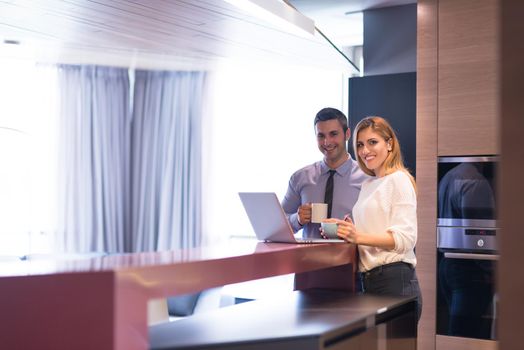 A young couple is preparing for the job and using a laptop. The man drinks coffee while the woman eats breakfast at luxury home together, looking at screen, smiling.