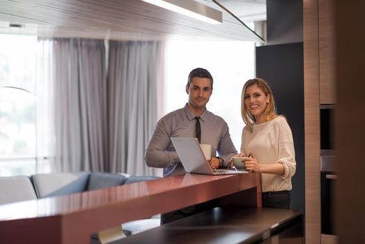 A young couple is preparing for the job and using a laptop. The man drinks coffee while the woman eats breakfast at luxury home together, looking at screen, smiling.