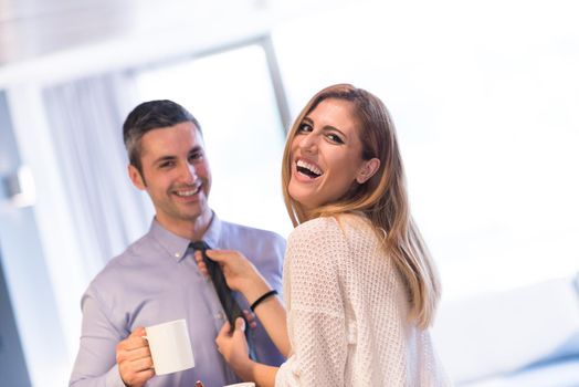 A young couple is preparing for the job and using a laptop. The man drinks coffee while the woman eats breakfast at luxury home together, looking at screen, smiling.