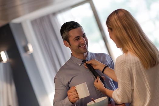 A young couple is preparing for the job and using a laptop. The man drinks coffee while the woman eats breakfast at luxury home together, looking at screen, smiling.