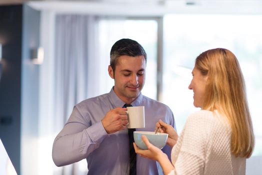 A young couple is preparing for the job and using a laptop. The man drinks coffee while the woman eats breakfast at luxury home together, looking at screen, smiling.