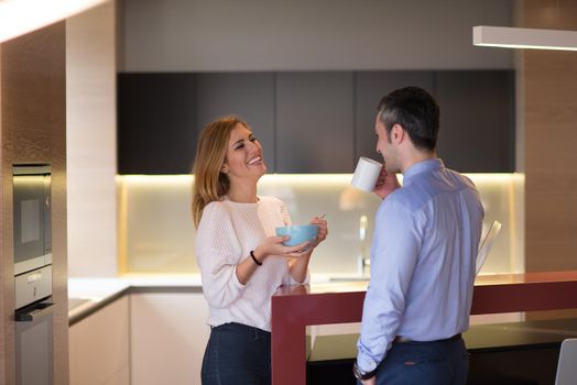 A young couple is preparing for the job and using a laptop. The man drinks coffee while the woman eats breakfast at luxury home together, looking at screen, smiling.