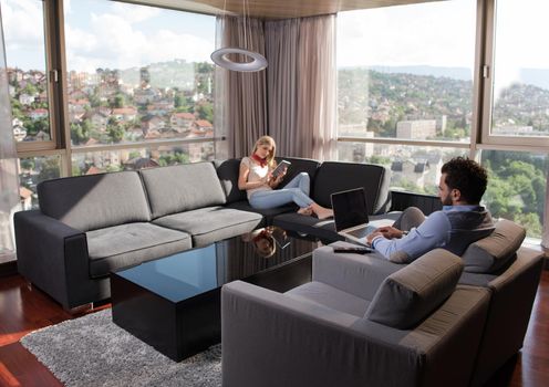 Young couple relaxing at home using tablet and laptop computers reading near the window on the sofa couch.