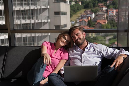 Young couple relaxing at  home using laptop computers reading in the living room near the window on the sofa couch.