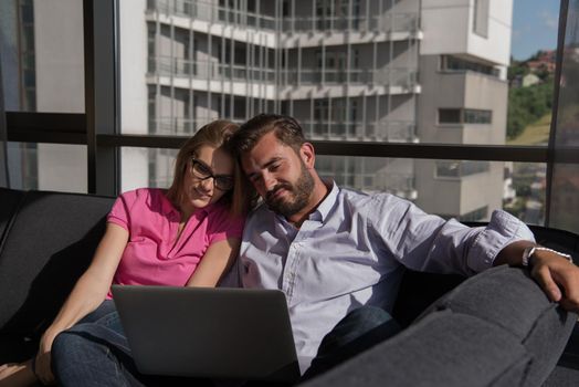 Young couple relaxing at  home using laptop computers reading in the living room near the window on the sofa couch.