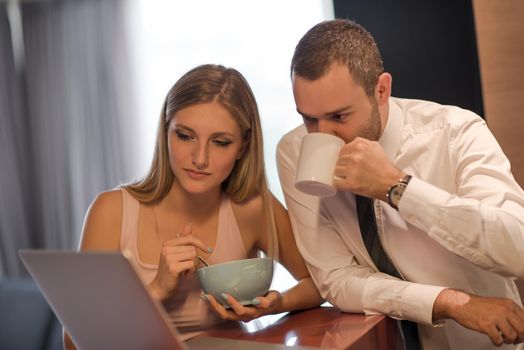 A young couple is preparing for the job and using a laptop. The man drinks coffee while the woman eats breakfast at luxury home together, looking at screen, smiling.