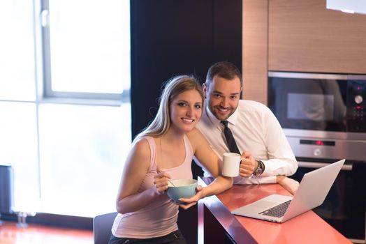 A young couple is preparing for the job and using a laptop. The man drinks coffee while the woman eats breakfast at luxury home together, looking at screen, smiling.