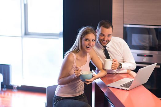 A young couple is preparing for the job and using a laptop. The man drinks coffee while the woman eats breakfast at luxury home together, looking at screen, smiling.