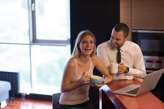 A young couple is preparing for the job and using a laptop. The man drinks coffee while the woman eats breakfast at luxury home together, looking at screen, smiling.