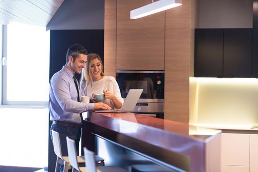 A young couple is preparing for the job and using a laptop. The man drinks coffee while the woman eats breakfast at luxury home together, looking at screen, smiling.