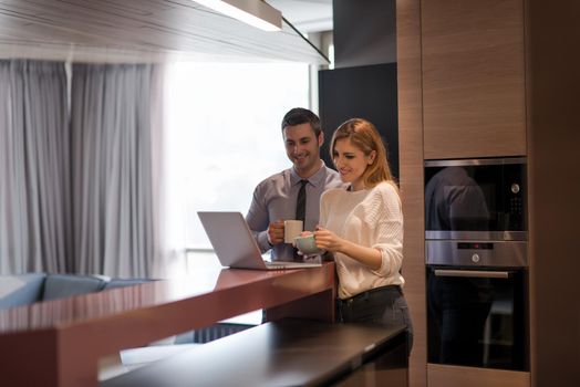 A young couple is preparing for the job and using a laptop. The man drinks coffee while the woman eats breakfast at luxury home together, looking at screen, smiling.