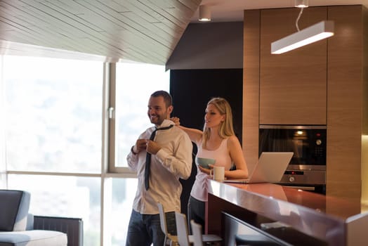 A young couple is preparing for the job and using a laptop. The man drinks coffee while the woman eats breakfast at luxury home together, looking at screen, smiling.