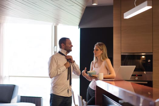 A young couple is preparing for the job and using a laptop. The man drinks coffee while the woman eats breakfast at luxury home together, looking at screen, smiling.