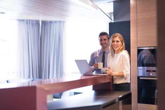 A young couple is preparing for the job and using a laptop. The man drinks coffee while the woman eats breakfast at luxury home together, looking at screen, smiling.