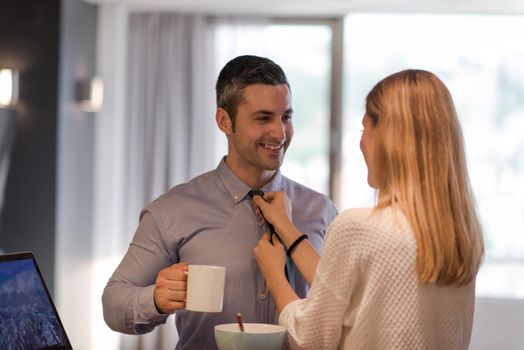 A young couple is preparing for the job and using a laptop. The man drinks coffee while the woman eats breakfast at luxury home together, looking at screen, smiling.