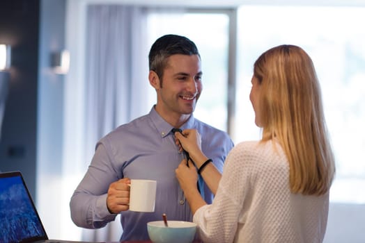 A young couple is preparing for the job and using a laptop. The man drinks coffee while the woman eats breakfast at luxury home together, looking at screen, smiling.