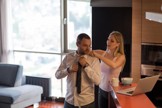 A young couple is preparing for the job and using a laptop. The man drinks coffee while the woman eats breakfast at luxury home together, looking at screen, smiling.