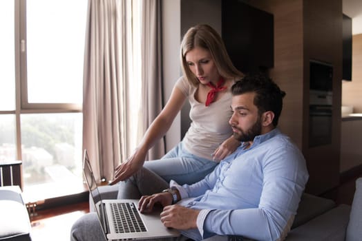 Young couple relaxing at  home using laptop computers reading in the living room near the window on the sofa couch.