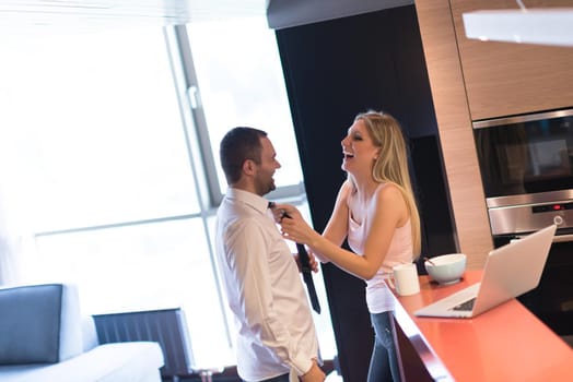 A young couple is preparing for the job and using a laptop. The man drinks coffee while the woman eats breakfast at luxury home together, looking at screen, smiling.