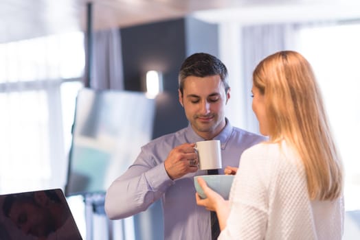 A young couple is preparing for the job and using a laptop. The man drinks coffee while the woman eats breakfast at luxury home together, looking at screen, smiling.