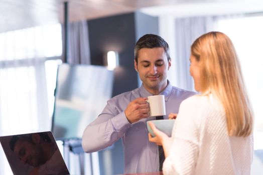 A young couple is preparing for the job and using a laptop. The man drinks coffee while the woman eats breakfast at luxury home together, looking at screen, smiling.