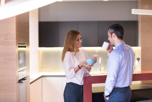 A young couple is preparing for the job and using a laptop. The man drinks coffee while the woman eats breakfast at luxury home together, looking at screen, smiling.