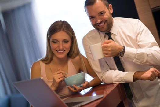 A young couple is preparing for the job and using a laptop. The man drinks coffee while the woman eats breakfast at luxury home together, looking at screen, smiling.