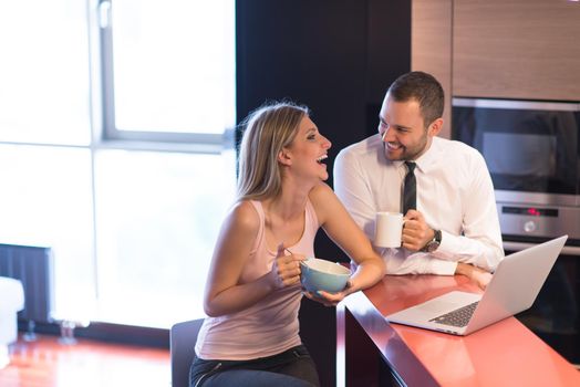 A young couple is preparing for the job and using a laptop. The man drinks coffee while the woman eats breakfast at luxury home together, looking at screen, smiling.