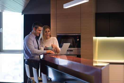 A young couple is preparing for the job and using a laptop. The man drinks coffee while the woman eats breakfast at luxury home together, looking at screen, smiling.