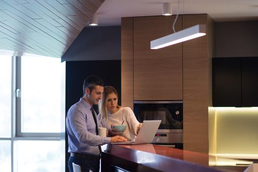 A young couple is preparing for the job and using a laptop. The man drinks coffee while the woman eats breakfast at luxury home together, looking at screen, smiling.