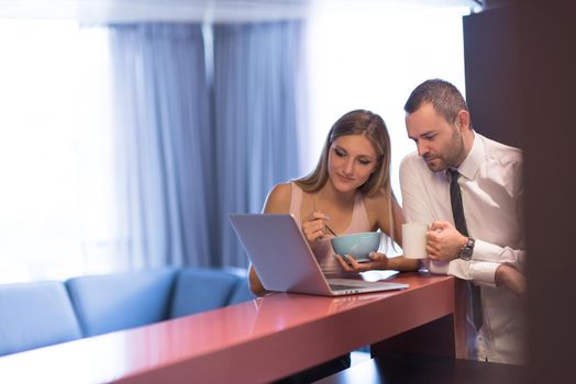 A young couple is preparing for the job and using a laptop. The man drinks coffee while the woman eats breakfast at luxury home together, looking at screen, smiling.