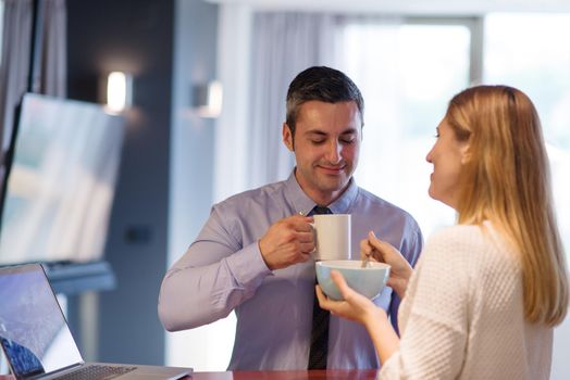 A young couple is preparing for the job and using a laptop. The man drinks coffee while the woman eats breakfast at luxury home together, looking at screen, smiling.