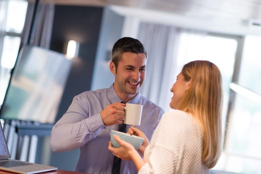 A young couple is preparing for the job and using a laptop. The man drinks coffee while the woman eats breakfast at luxury home together, looking at screen, smiling.