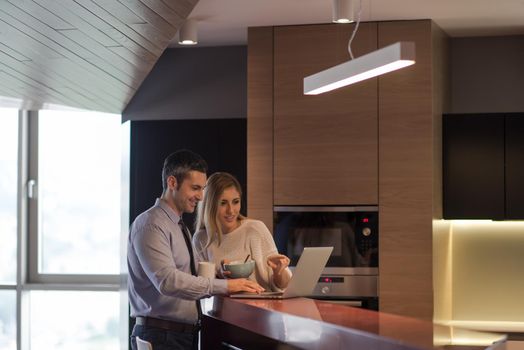 A young couple is preparing for the job and using a laptop. The man drinks coffee while the woman eats breakfast at luxury home together, looking at screen, smiling.