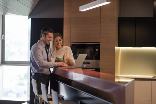 A young couple is preparing for the job and using a laptop. The man drinks coffee while the woman eats breakfast at luxury home together, looking at screen, smiling.