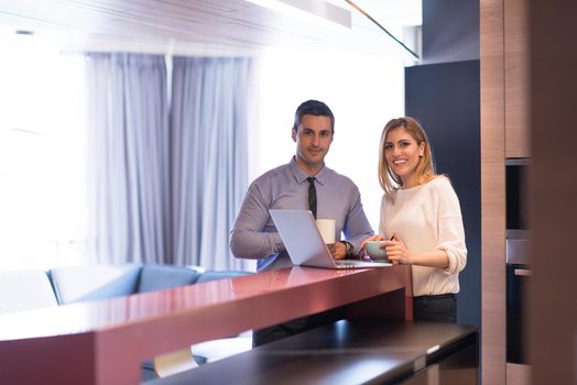 A young couple is preparing for the job and using a laptop. The man drinks coffee while the woman eats breakfast at luxury home together, looking at screen, smiling.