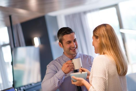 A young couple is preparing for the job and using a laptop. The man drinks coffee while the woman eats breakfast at luxury home together, looking at screen, smiling.