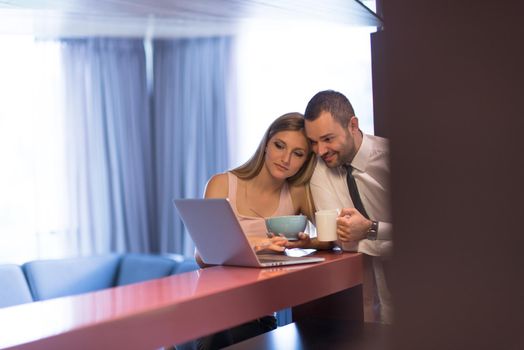 A young couple is preparing for the job and using a laptop. The man drinks coffee while the woman eats breakfast at luxury home together, looking at screen, smiling.