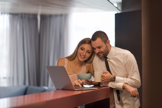 A young couple is preparing for the job and using a laptop. The man drinks coffee while the woman eats breakfast at luxury home together, looking at screen, smiling.