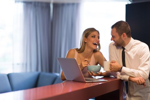 A young couple is preparing for the job and using a laptop. The man drinks coffee while the woman eats breakfast at luxury home together, looking at screen, smiling.