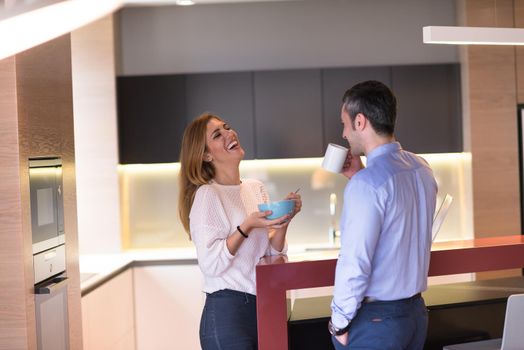 A young couple is preparing for the job and using a laptop. The man drinks coffee while the woman eats breakfast at luxury home together, looking at screen, smiling.