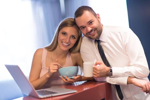 A young couple is preparing for the job and using a laptop. The man drinks coffee while the woman eats breakfast at luxury home together, looking at screen, smiling.