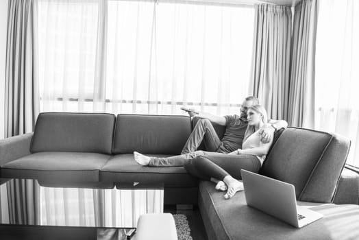 Young couple relaxes on the sofa in the luxury living room, using a laptop and remote control