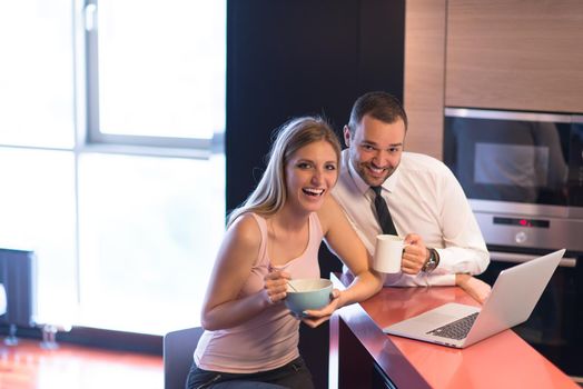 A young couple is preparing for the job and using a laptop. The man drinks coffee while the woman eats breakfast at luxury home together, looking at screen, smiling.