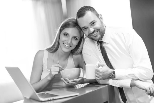 A young couple is preparing for the job and using a laptop. The man drinks coffee while the woman eats breakfast at luxury home together, looking at screen, smiling.