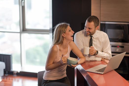 A young couple is preparing for the job and using a laptop. The man drinks coffee while the woman eats breakfast at luxury home together, looking at screen, smiling.