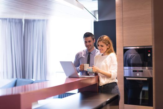 A young couple is preparing for the job and using a laptop. The man drinks coffee while the woman eats breakfast at luxury home together, looking at screen, smiling.