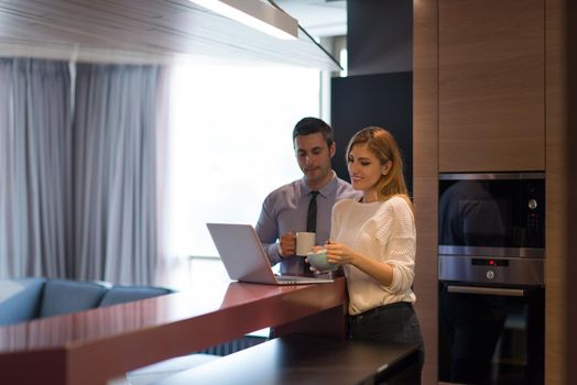 A young couple is preparing for the job and using a laptop. The man drinks coffee while the woman eats breakfast at luxury home together, looking at screen, smiling.