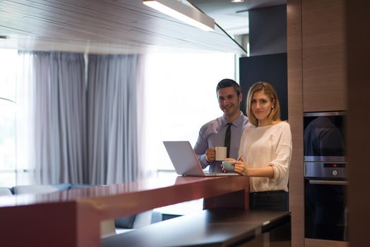 A young couple is preparing for the job and using a laptop. The man drinks coffee while the woman eats breakfast at luxury home together, looking at screen, smiling.