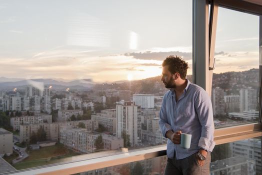 young man enjoying evening coffee and beautiful sunset landscape of the city while standing by the window